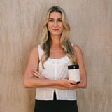 A blonde woman stands against a textured beige wall, wearing a white sleeveless blouse and black pants while holding a white jar with a black lid.