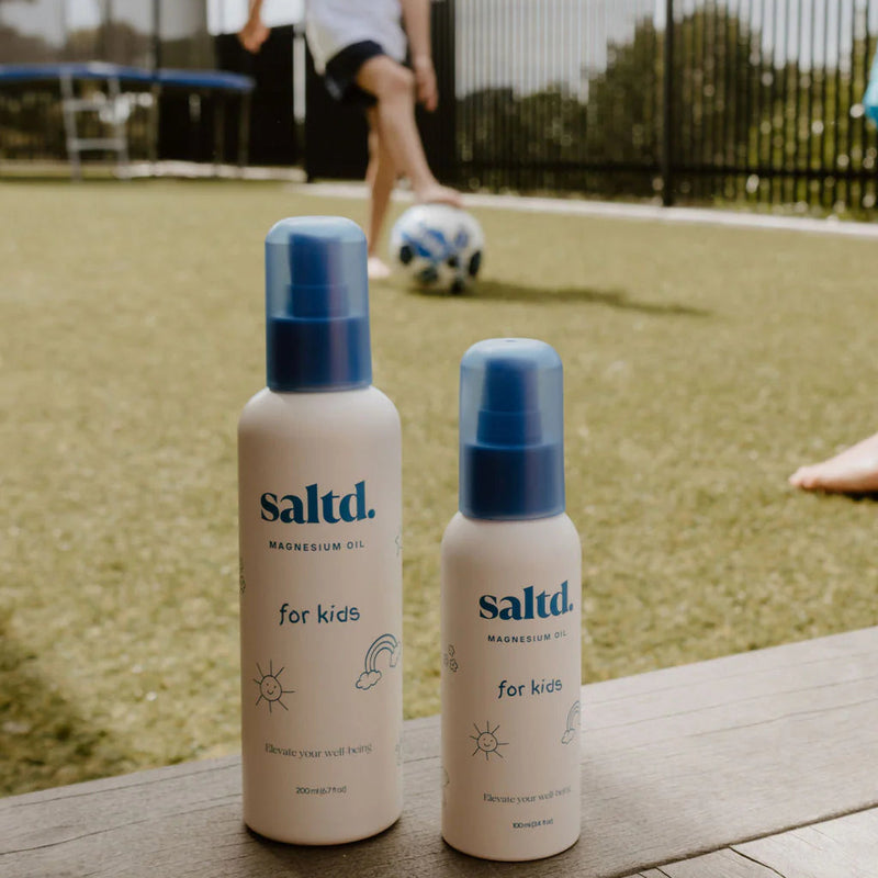 Two bottles of saltd product for kids on a wooden surface with children playing in the background.