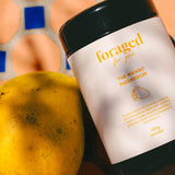 A black plastic tub of 'foraged for you' The Mango Magnesium powder, styled next to a fresh yellow mango on a blue and terracotta mosaic tile background.