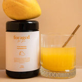 A jar of Foraged For You The Mango Magnesium powder sits next to a glass of orange-yellow liquid with a metal spoon, with a fresh mango resting on top of the jar, all set against a warm beige background.